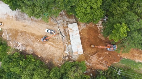 Aerial picture of road being repaired with cranes and trucks. Trees line each side of the road