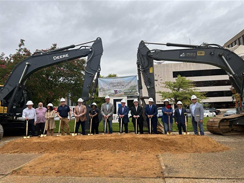 Group of people in business attire and hard hats, holding shovels and standing in front of a small dirt mound, flanking the arms of two large excavators, for a 