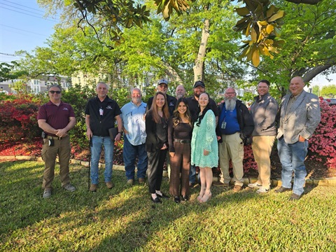 Group of twelve people, men and women, smiling and posing for a photo outdoors in front of a blooming azalea bush, with foliage and sky in the background.