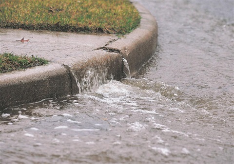 Heavy rainfall overflowing a concrete curb, with water cascading into a flooded street; a small patch of grass sits atop the curb.