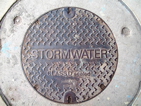 Close-up of a circular, dark metal storm drain cover set in concrete. The cover is patterned with rows of angled chevrons, and embossed with the word 