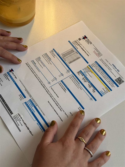 A person's hands with gold painted nails holding a City of Tyler utilities bill on a white surface, with part of a clear glass of liquid at the upper left corner of the image.